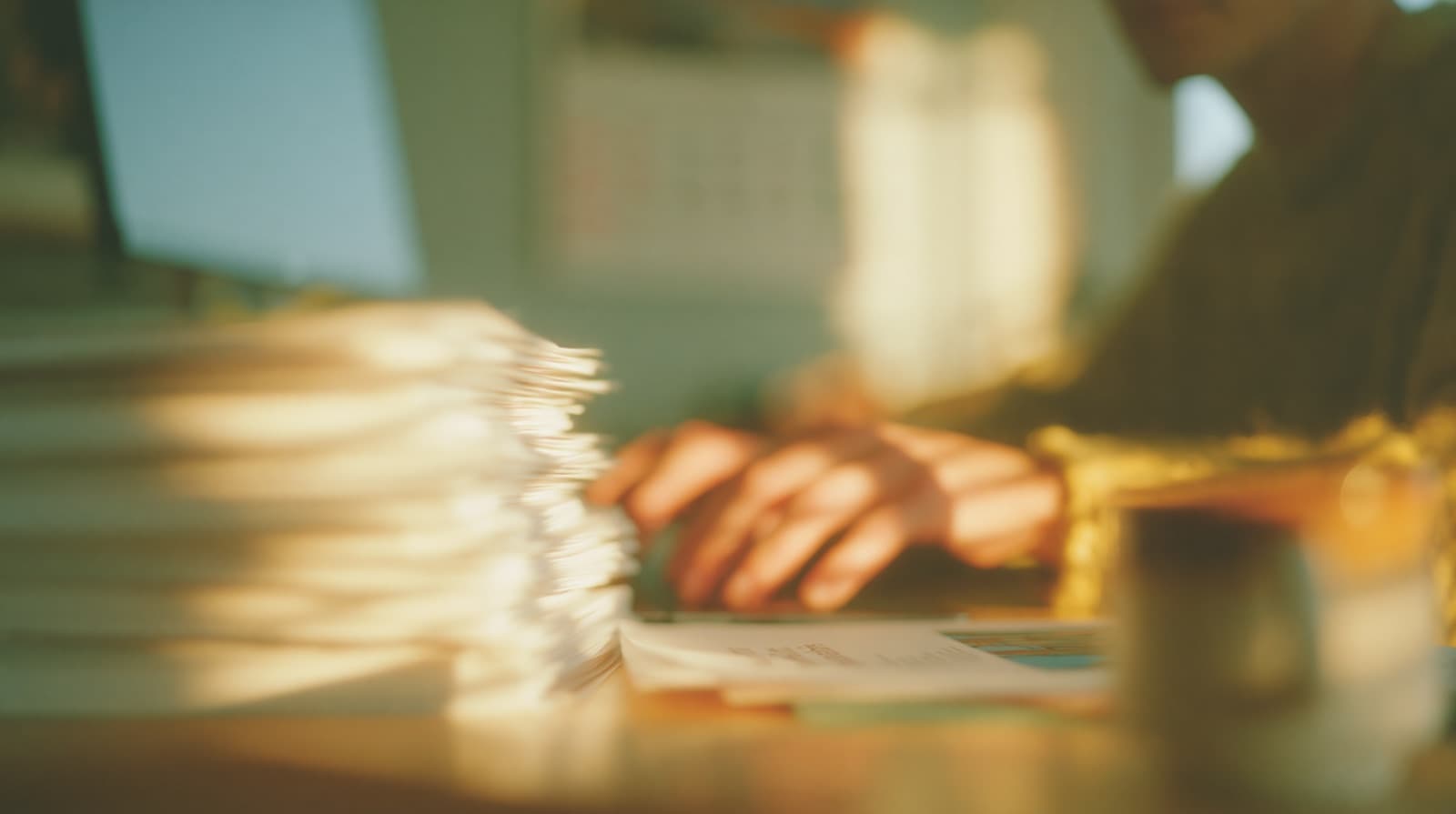 Hands at a desk beside a tall stack of printed reports in warm light