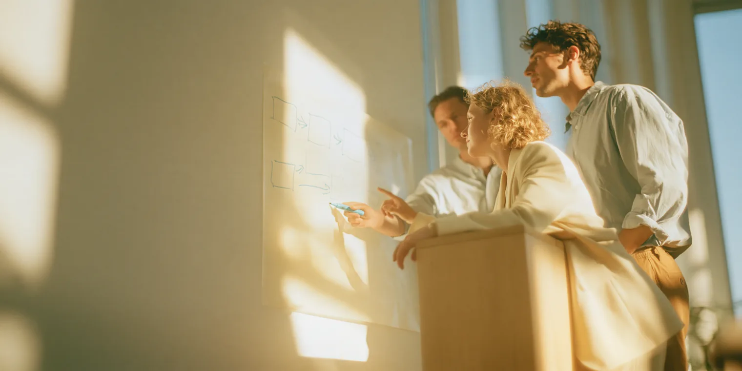 Three colleagues at a light-wood counter in a sunlit office, one tracing a hand-drawn workflow diagram on a whiteboard